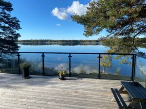 Terrasse en bois avec garde-corps à poteaux noirs encadrant une propriété au bord du lac, offrant une vue dégagée sur l’eau et la nature.