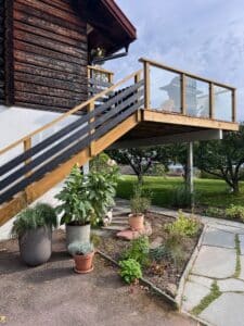 Terrasse surélevée avec garde-corps en bois et verre clair contre une façade en rondins, vue sur jardin et végétation.