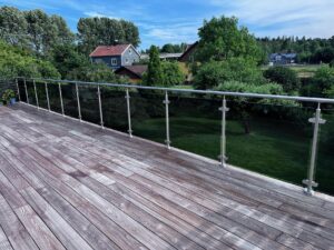 Garde-corps en verre avec main courante en acier inoxydable installé sur une terrasse en bois avec vue sur un jardin verdoyant.