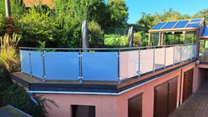 Terrasse avec garde-corps en verre dépoli et poteaux ronds en acier inoxydable – jardin et véranda en arrière-plan.