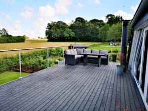 Terrasse avec garde-corps en acier inoxydable et câbles, platelage gris et coin salon avec vue sur champs et campagne.