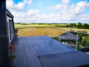 Garde-corps inox à câbles sur terrasse en bois gris foncé, vue dégagée sur champs et verdure environnante.