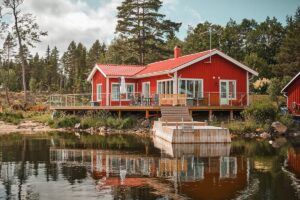 Garde-corps en verre installé autour d’une maison rouge au bord d’un lac, avec terrasse en bois sur pilotis.