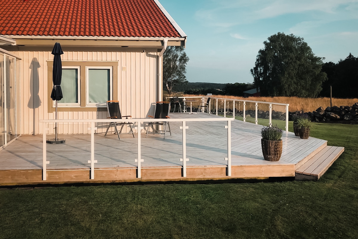 Balustrade blanche en aluminium et verre clair entourant vaste terrasse bois d’une maison campagnarde au toit rouge.
