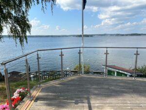 Terrasse avec garde-corps en verre et poteaux ronds en inox – vue sur lac paisible et passerelle en bois.