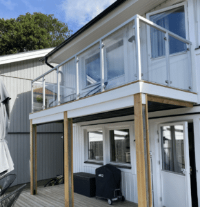 Garde-corps en verre avec montants blancs sur un balcon en bois, installé sur une maison moderne à deux étages.