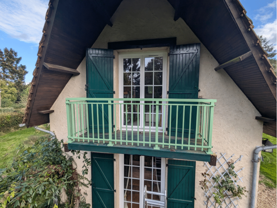 Garde-corps en aluminium vert clair au design classique, installé sur un petit balcon contre une façade enduite avec volets verts et charpente visible.