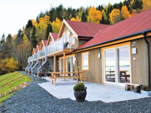 Rangée de chalets en bois avec garde-corps en verre sur les balcons, entourés de forêt aux couleurs d’automne.