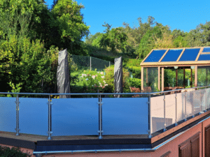 Garde-corps de balcon en acier inoxydable avec verre dépoli, monté sur terrasse surélevée. Poteaux ronds et main courante métallique. En fond : jardin verdoyant, serre vitrée et ciel bleu.