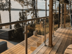 Garde-corps en verre avec poteaux inox et main courante en bois sur terrasse en bois. Panneaux maintenus par pinces. Vue sur lac entouré de forêt et escalier descendant vers l’eau.
