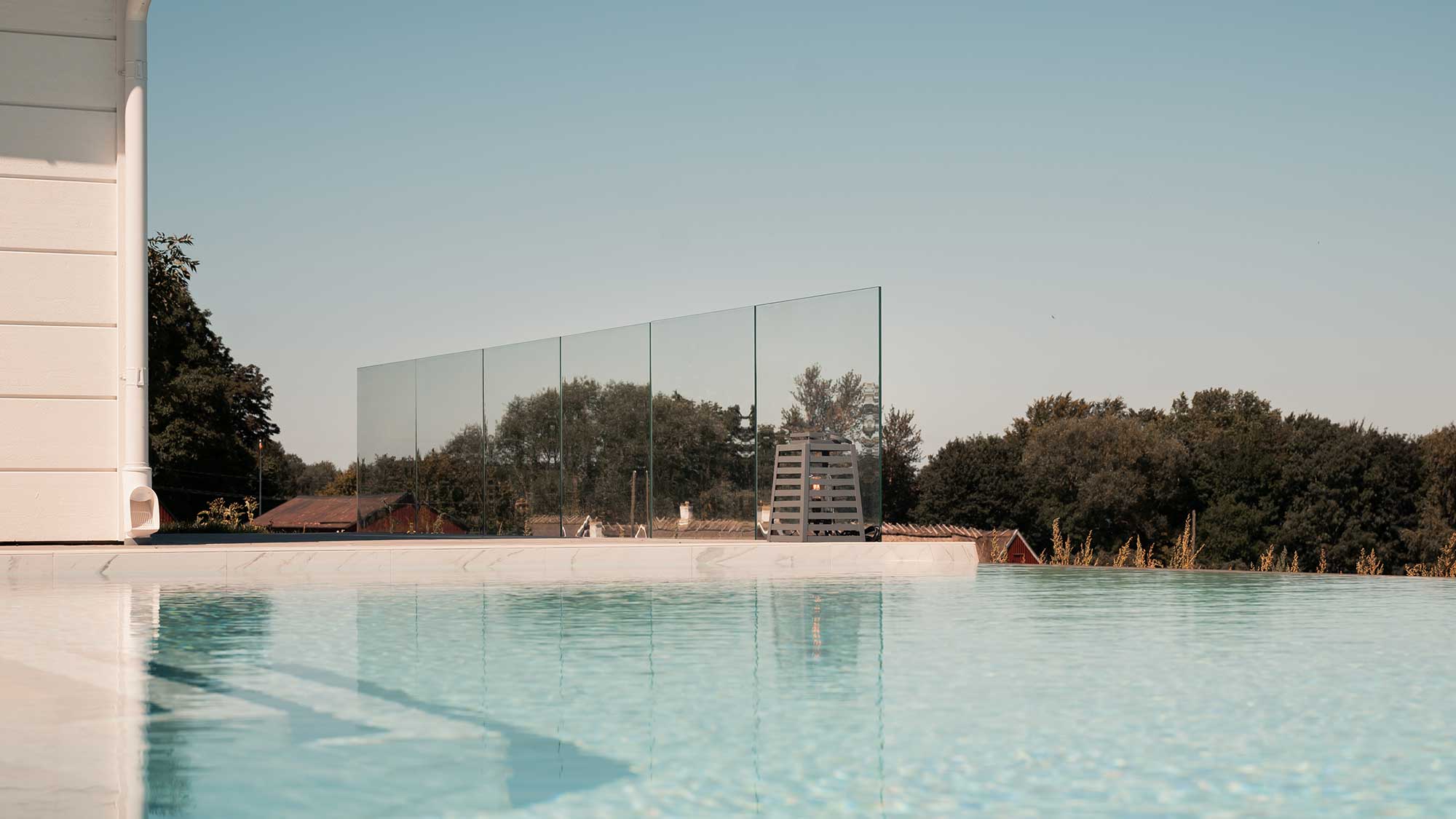 Garde-corps en verre sans poteaux monté latéralement près d’une piscine, avec vue dégagée sur des arbres et la mer en arrière-plan.